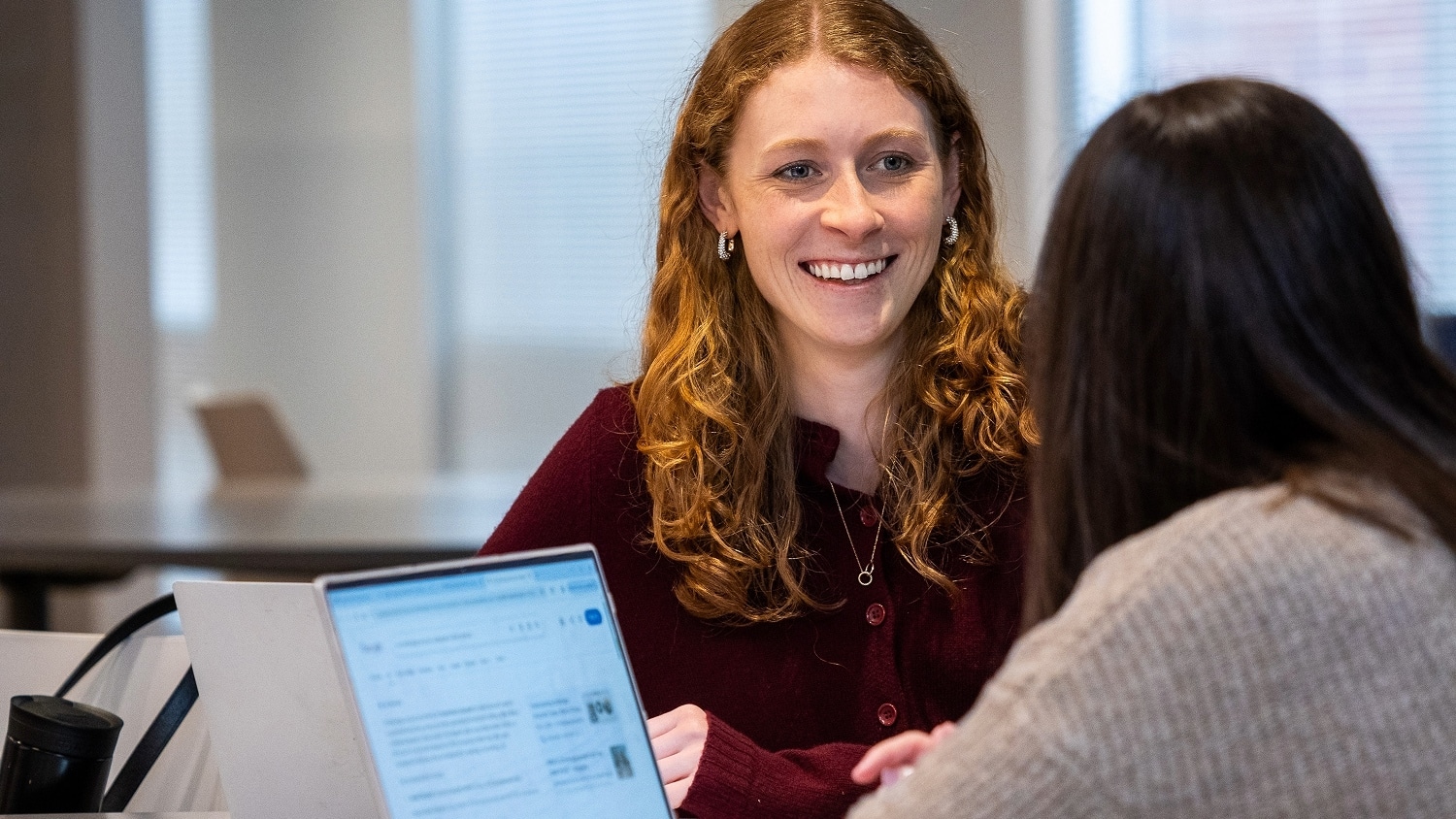 A woman smiling and engaging in conversation with a colleague while both work on laptops.