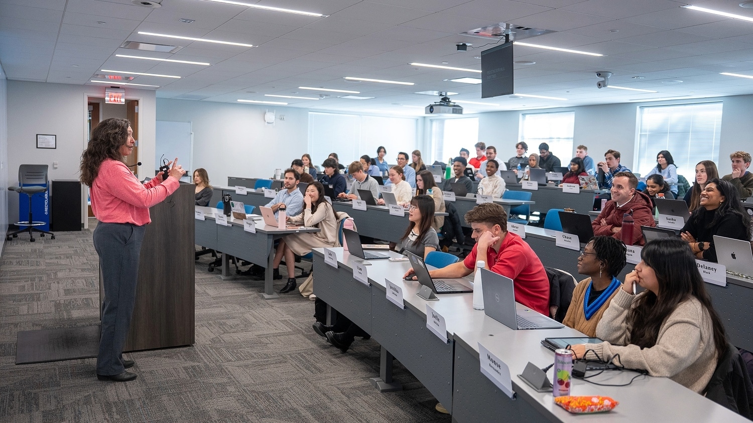 A professor speaks on stage during a lecture while many students sit in rows, listening attentively.