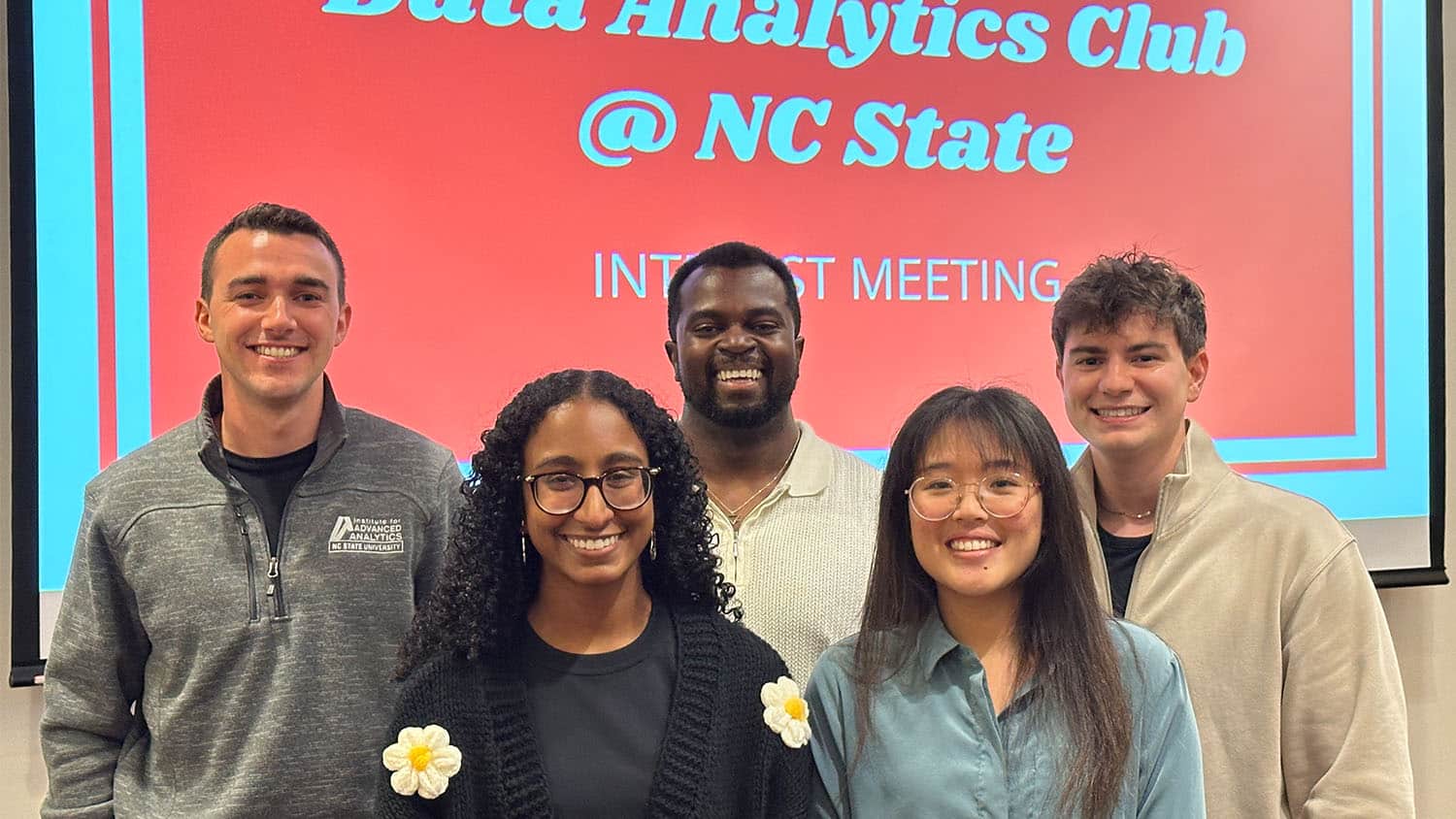 five alumni dressed in business casual attire stand together in front of a projector screen