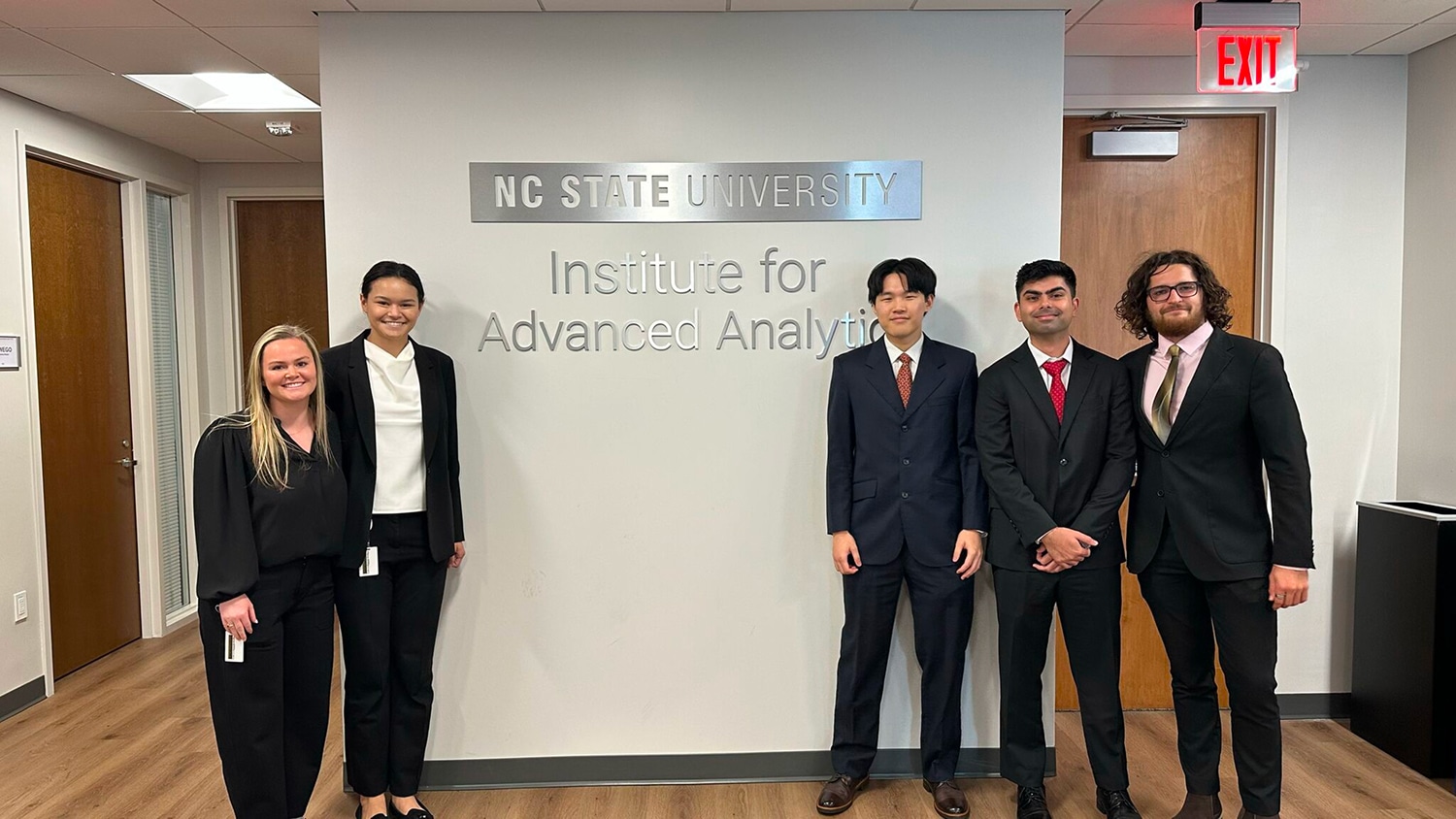 Five students in formal attire standing beside the IAA office sign.